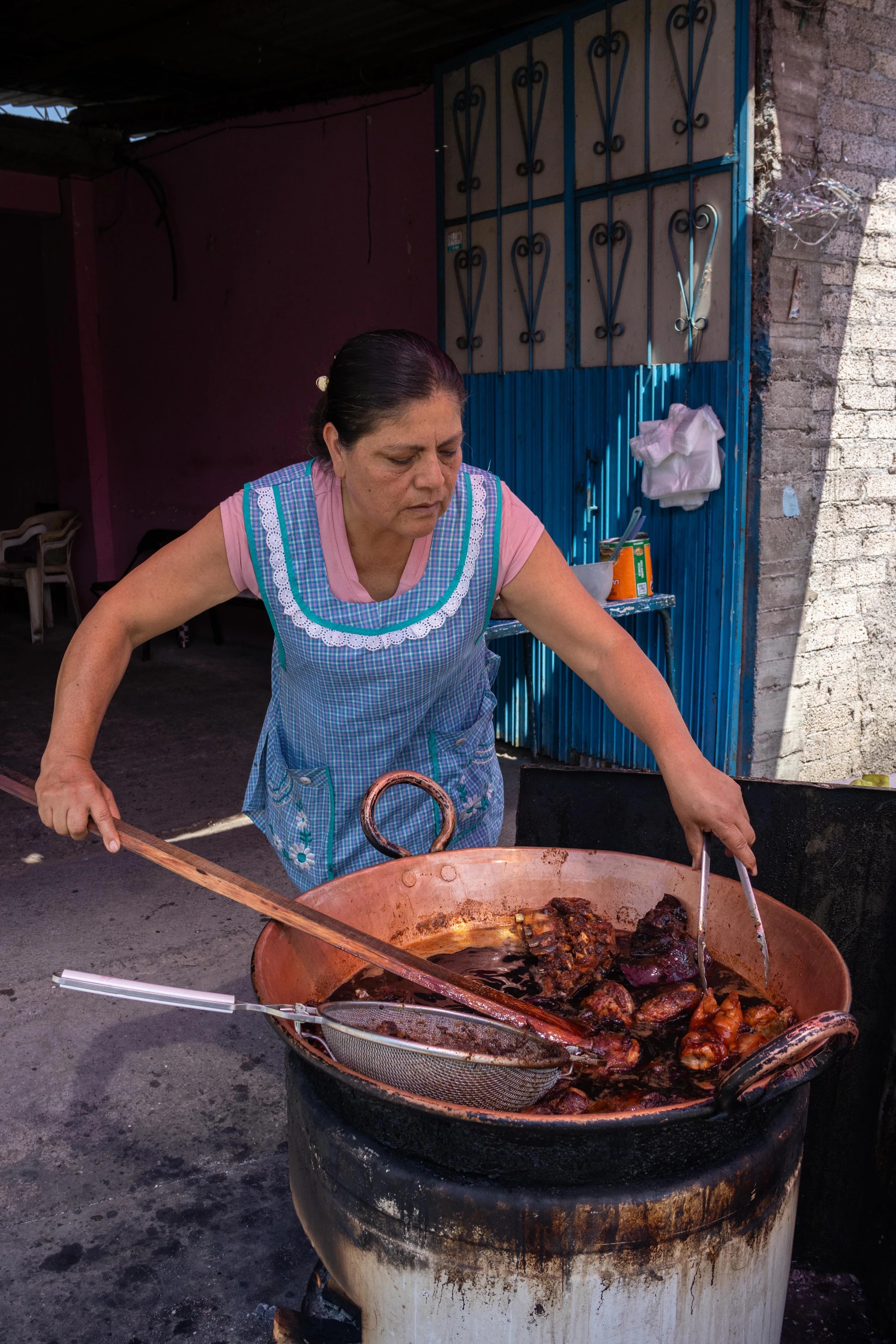 Teresa prepara carnitas con una pala de madera y unas pinzas en un cazo de cobre frente a una puerta azul