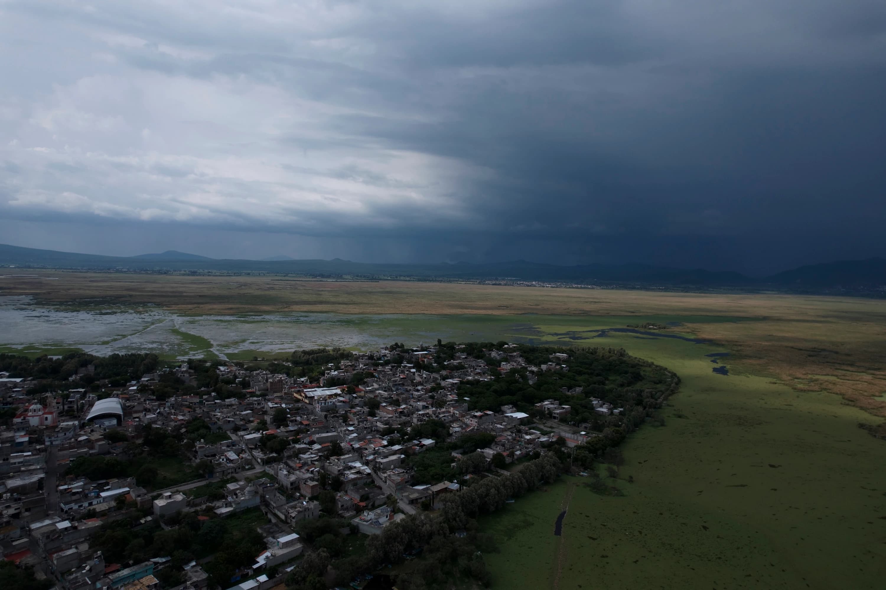 Vista aérea de Mariano Escobedo junto a los humedales del lago de Cuitzeo, con montañas al fondo y un cielo de nubes de tormenta