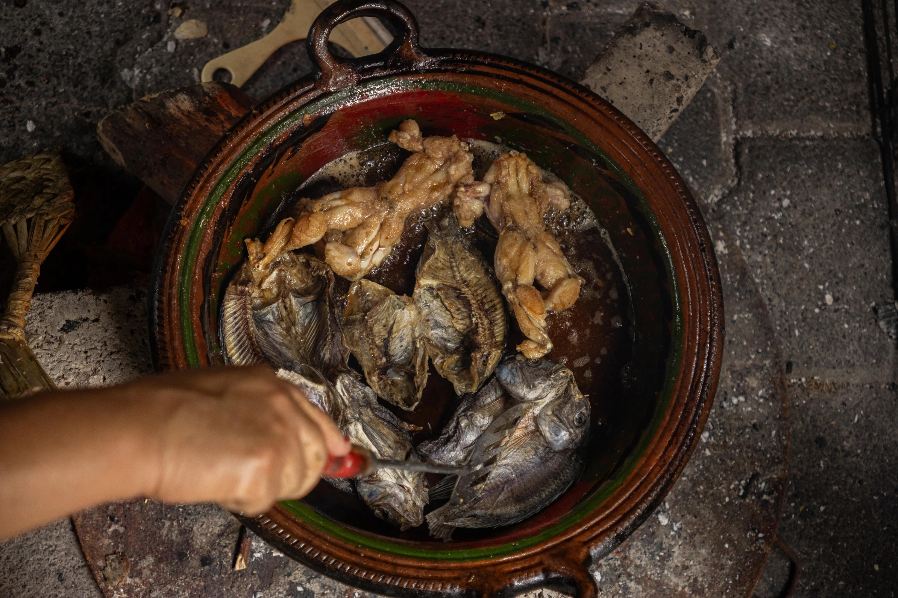 Ranas y pescado friéndose en una cazuela de barro mientras una mano los acomoda con un utensilio