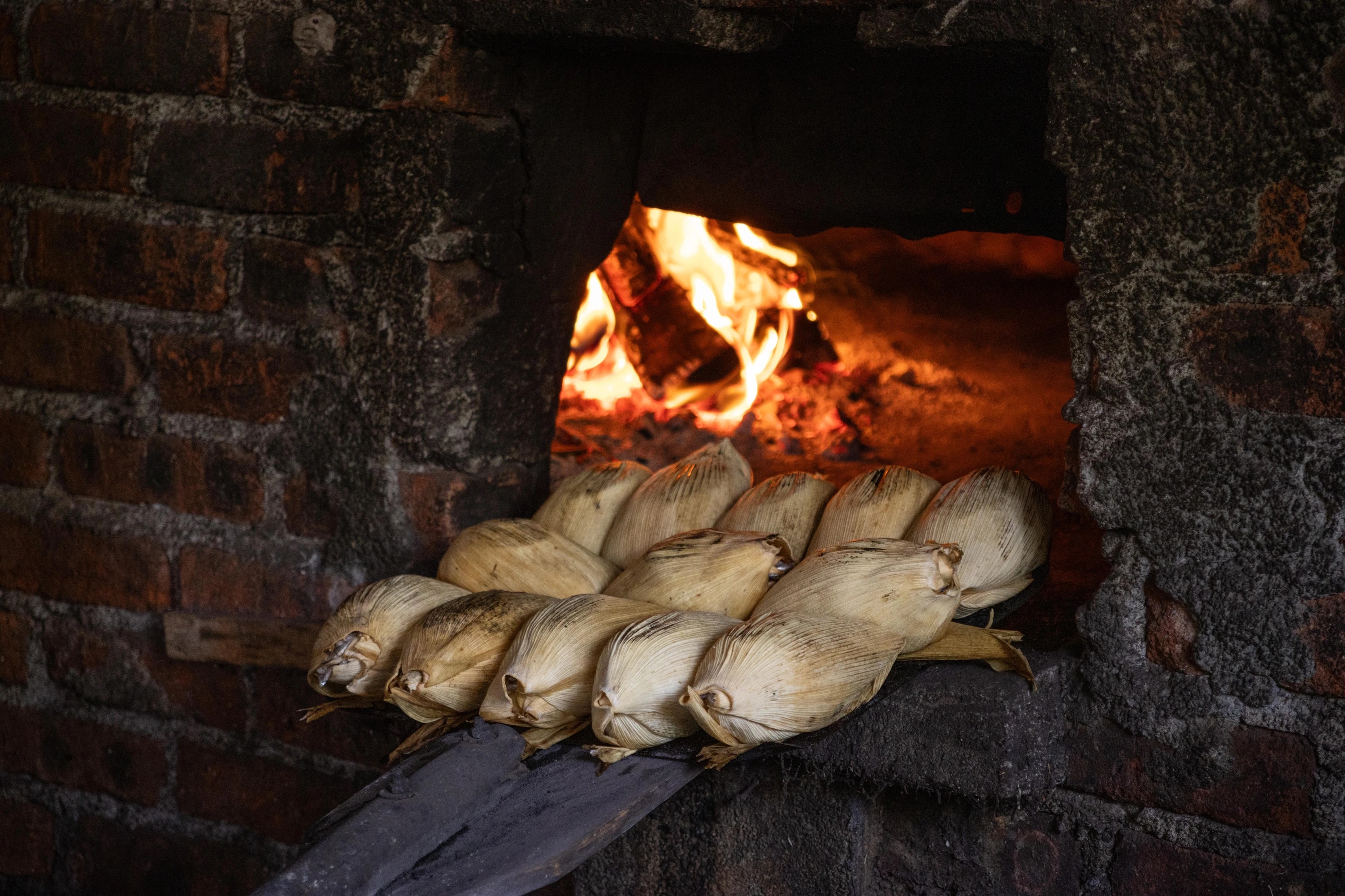 Tamales envueltos en hojas de maíz sobre una pala frente al fuego de un horno de ladrillo