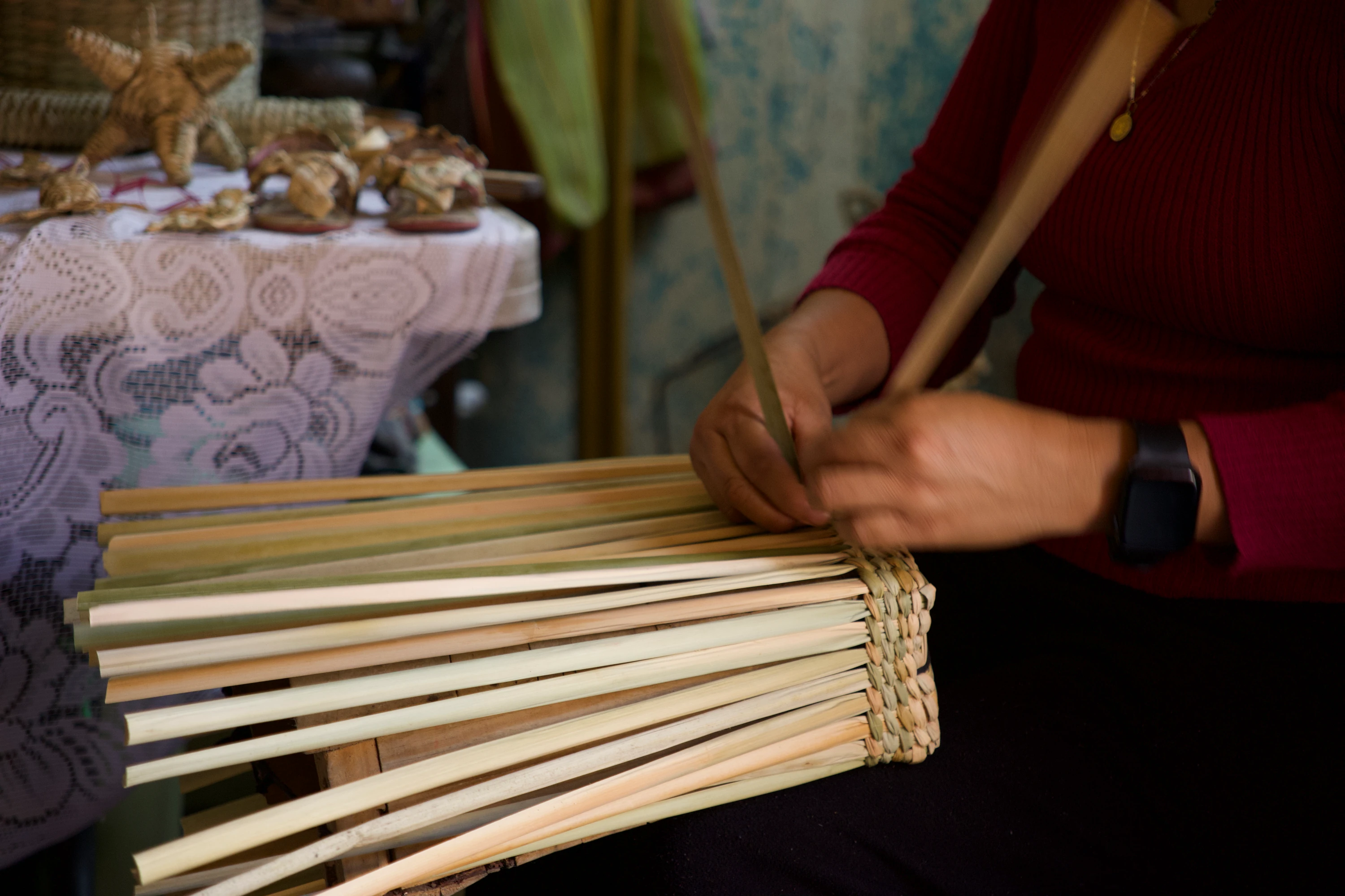 Manos de Antonia tejiendo tiras de tule, con piezas de artesanía sobre una mesa al fondo